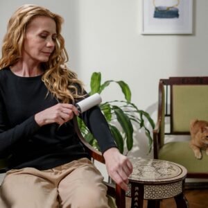 A woman cleaning her clothes with a lint roller while a ginger cat sits nearby, indoors.